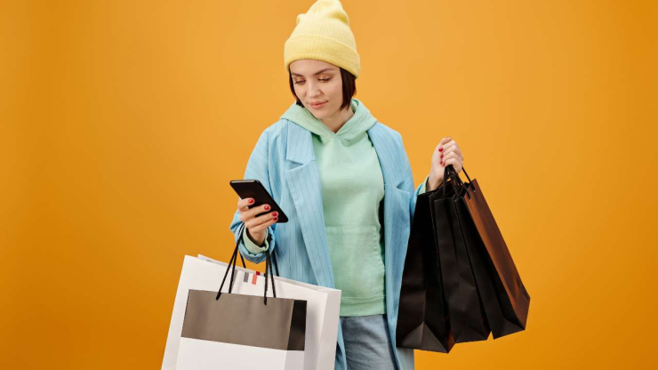 На картинке девушка в шапке держит сумки для покупок и телефон, а котором ищет мгновенный кредит